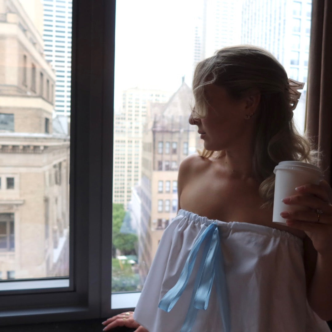 Woman in a white off-shoulder dress with a blue ribbon holding a coffee cup by a window.