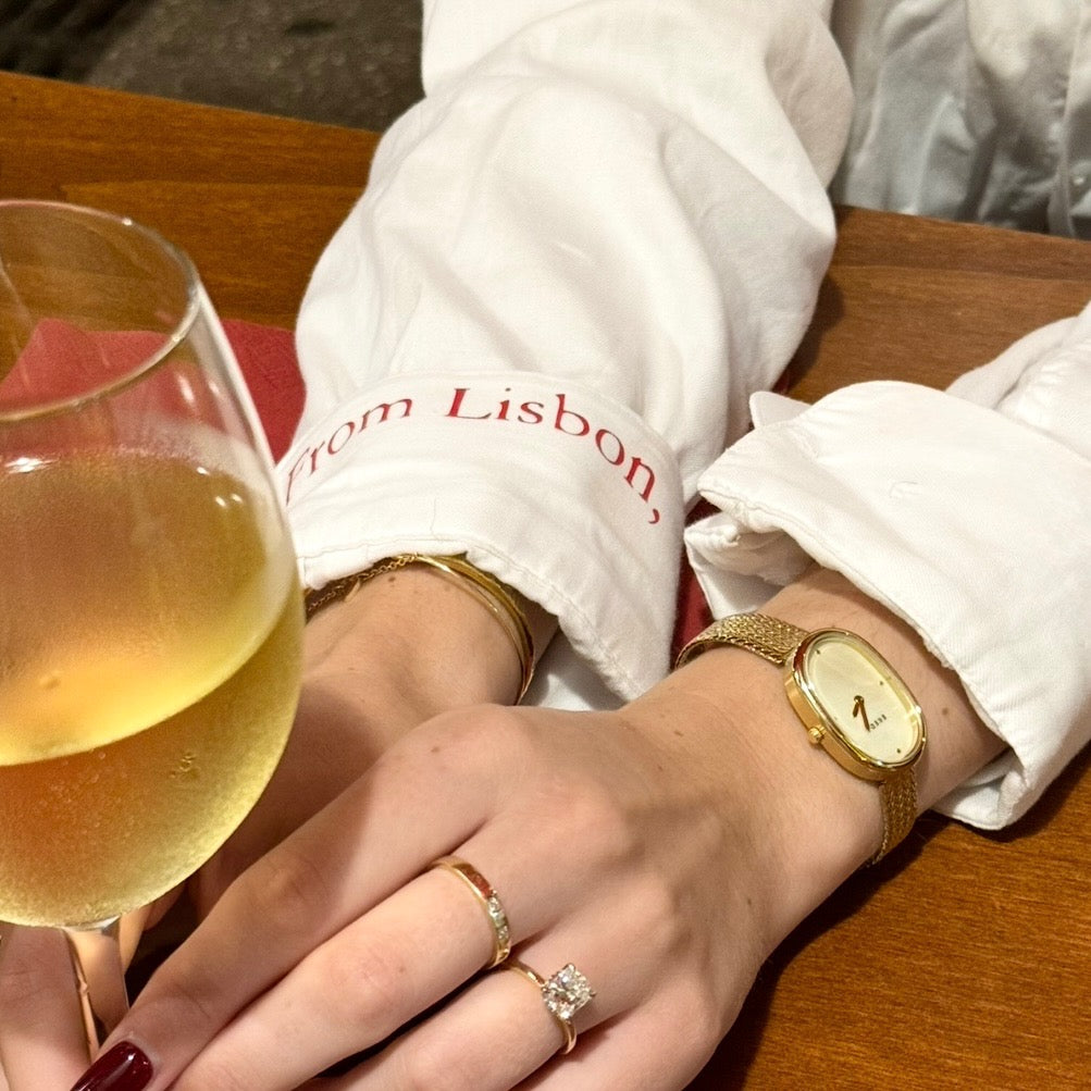 Person wearing a gold watch and ring, holding a glass of white wine, with a white shirt with 'from Lisbon' text.