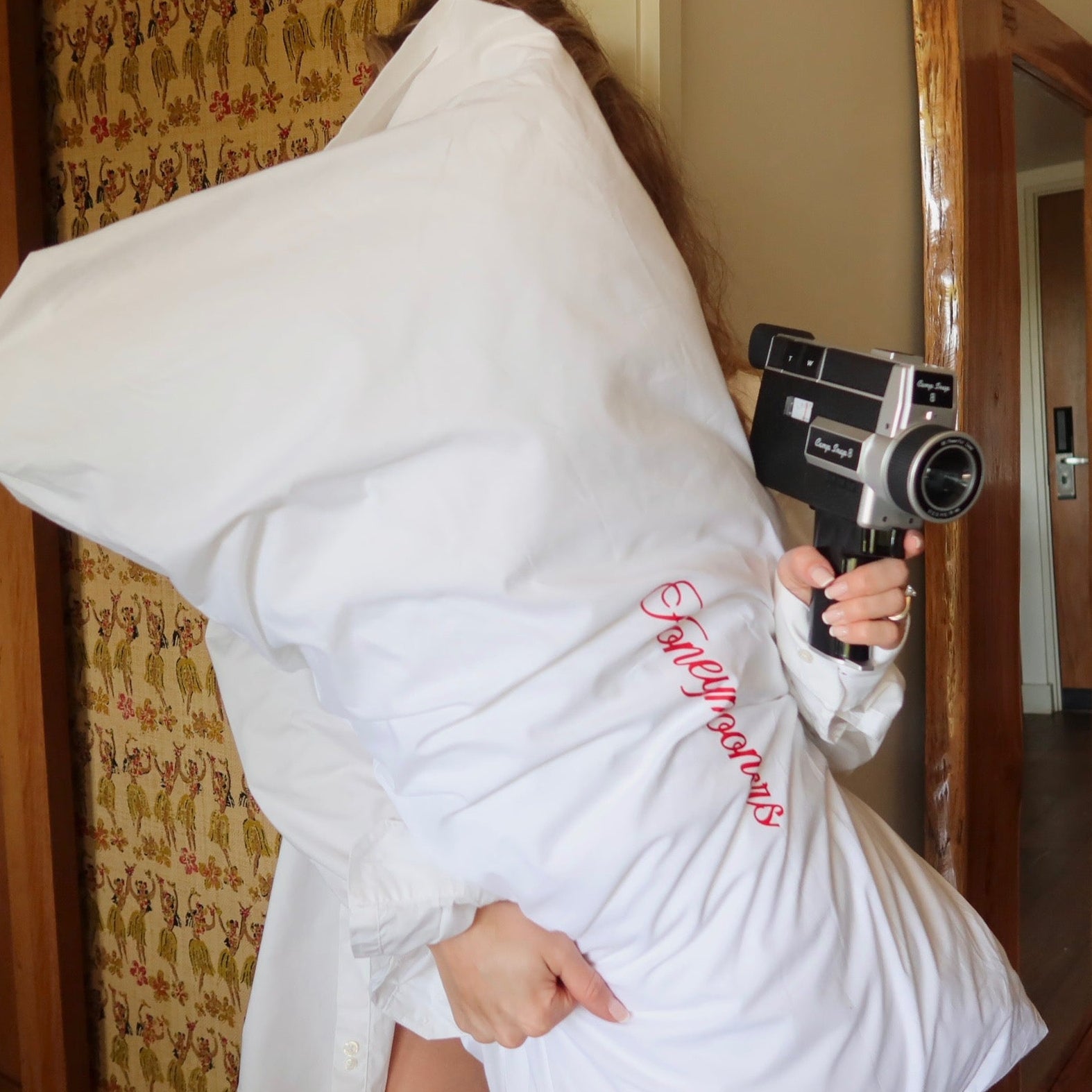 Person wrapped in a towel holding a vintage camera in a room with wooden doors and patterned wallpaper.